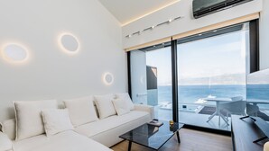 A modern living room with a white sofa, a glass coffee table, and a view of the sea through large windows. - GM Luxury Suites & Spa (Sitia)