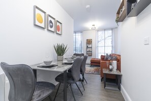 A modern living room with a dining area, featuring a sofa, a coffee table, and a dining table with chairs. - Elliot Oliver - Luxury 2 Bedroom Regency Apartment With Parking & EV Charger (Cheltenham)