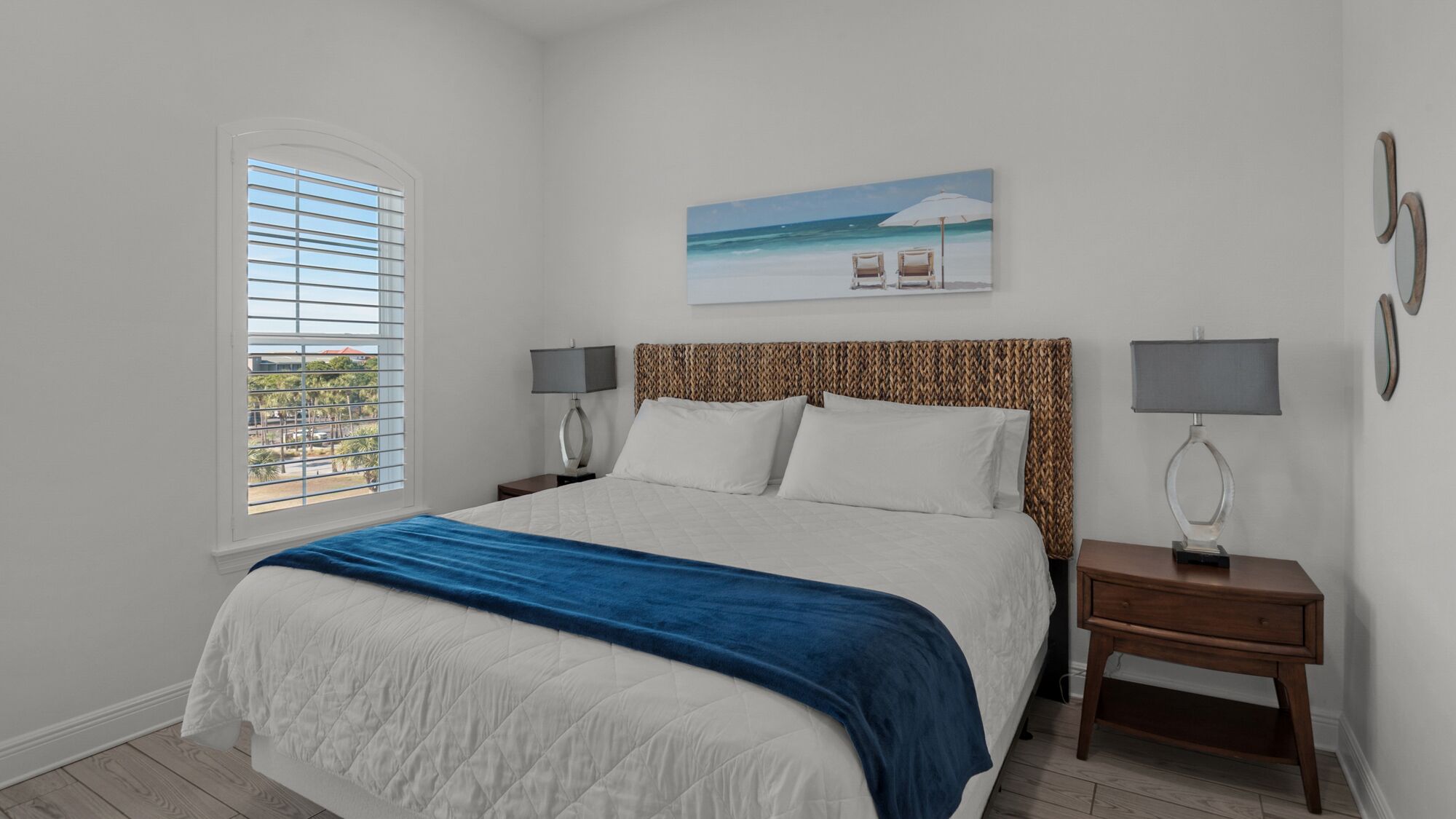 Un dormitorio con una cama, dos mesitas de noche con lámparas, una ventana con persianas y un cuadro de una playa.