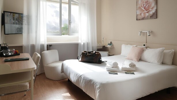 Een hotelkamer met een bed, een bureau, een stoel, een raam met gordijnen, een schilderij en een nachtkastje met lamp.