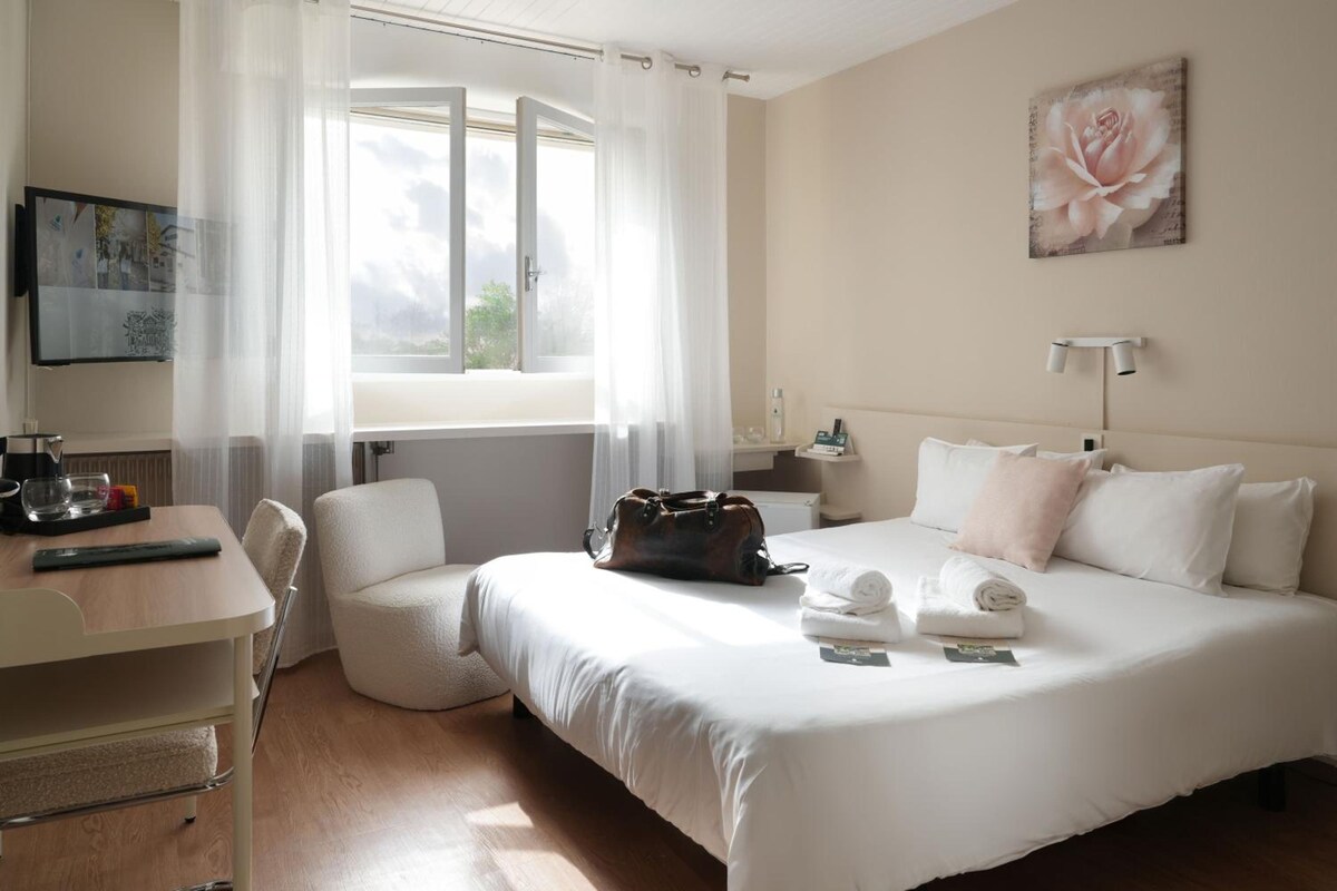 A hotel room with a bed, a desk, a chair, a window with curtains, a painting, and a nightstand with a lamp.