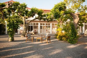 An outdoor seating area with tables and chairs, surrounded by trees and a building with large windows. - Logis Hotel Thermal (Saubusse)