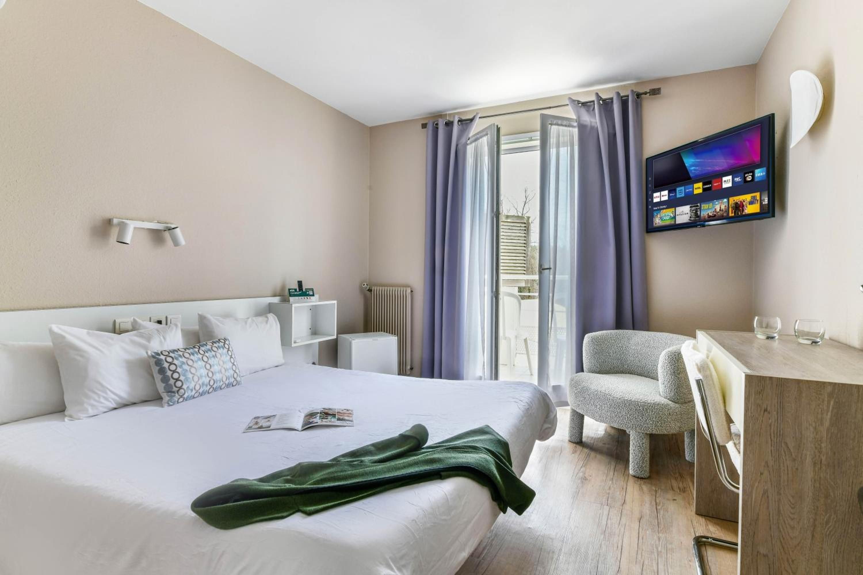 Quarto de hotel com cama, escrivaninha, cadeira, televisão e janela com cortinas.