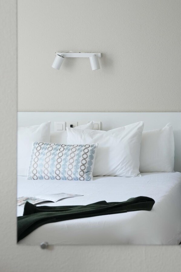 A neatly made bed with white bedding and a patterned pillow, a white bedside table, and a wall-mounted light fixture. - Logis Hotel Thermal (Saubusse)