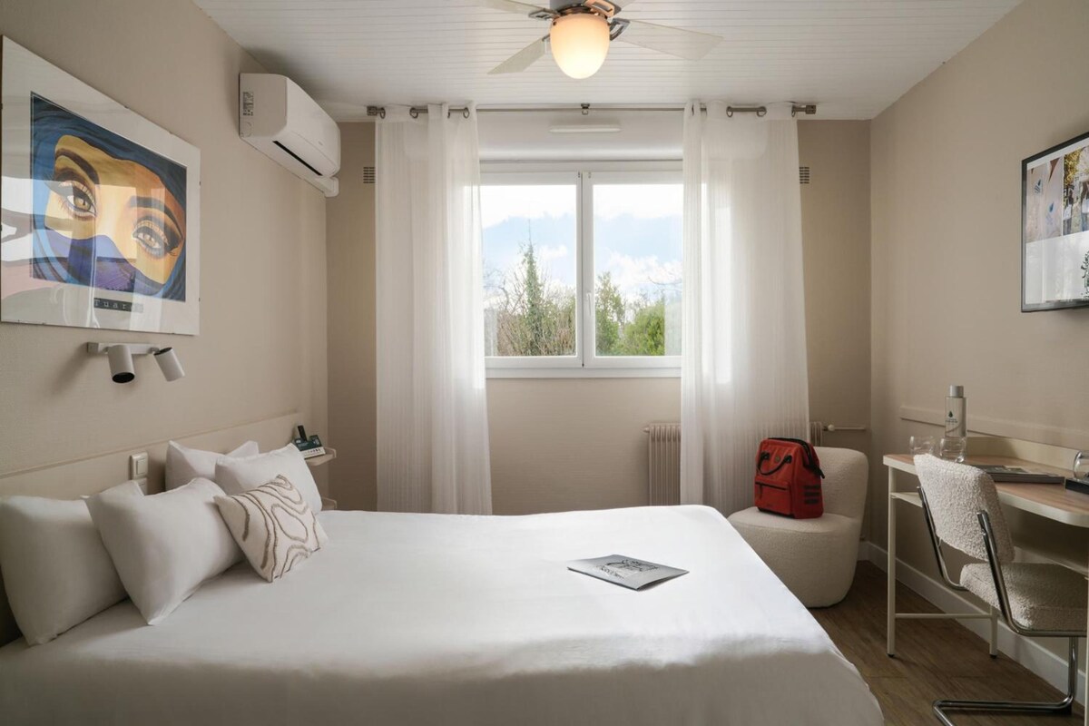 A hotel room with a bed, a desk, a chair, a window with curtains, and artwork on the walls.