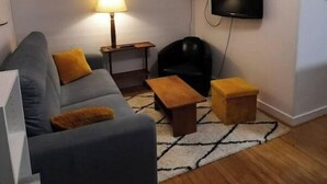 Un salón con un sofá gris, dos mesitas de madera, una lámpara de pie, un sillón negro y un televisor montado en la pared.