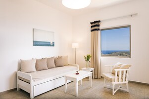 Une pièce au décor minimaliste, avec un canapé blanc, une petite table, une chaise, une lampe et une fenêtre donnant sur la mer.