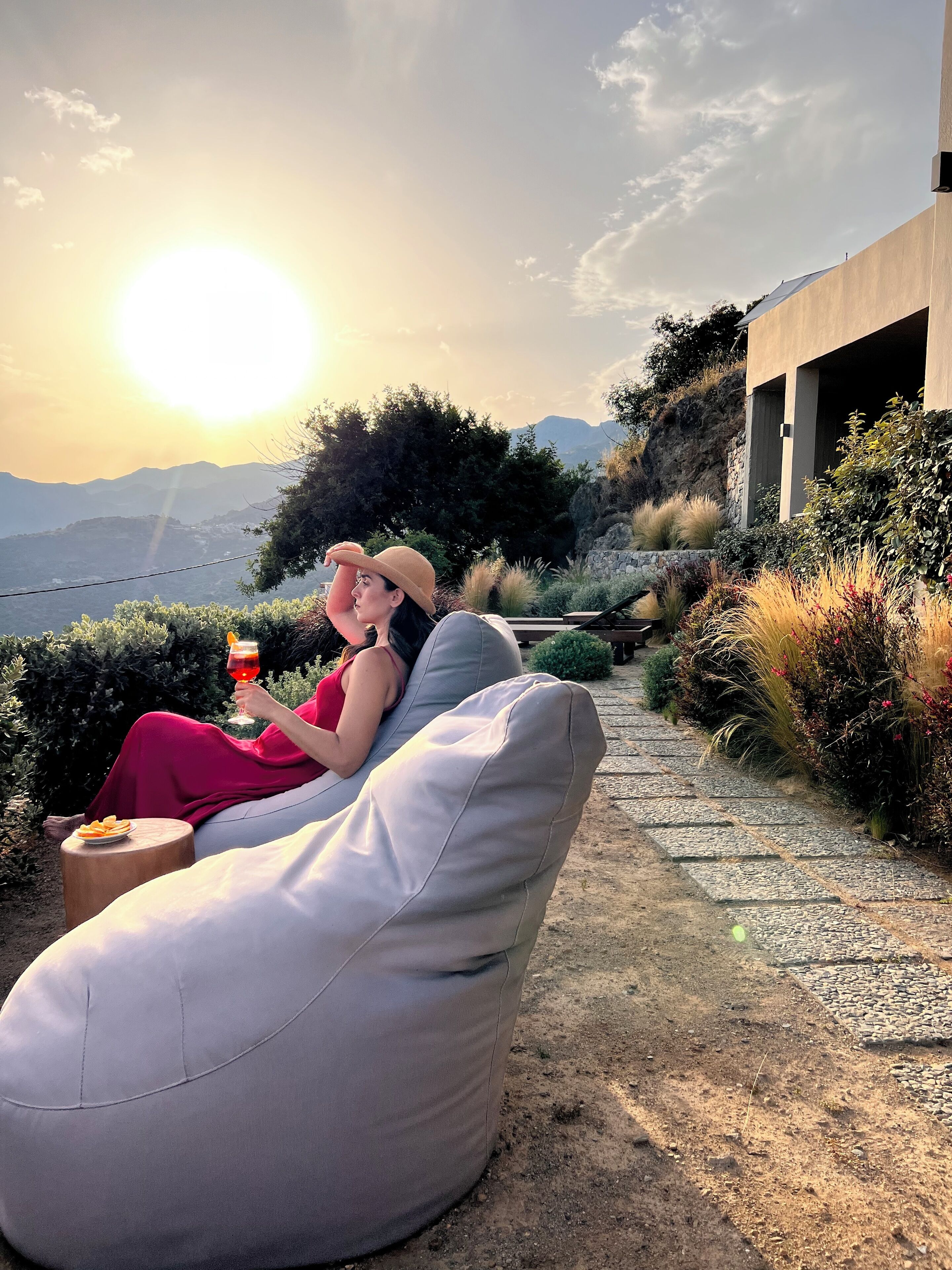 Eine Person entspannt auf einem beigen Outdoor-Sofa mit einem Getränk, umgeben von Grünpflanzen und einem Kiesweg, mit Bergen im Hintergrund.