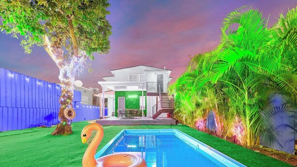 A swimming pool with two swan-shaped inflatable rings, surrounded by lush greenery and a house with a green wall and a staircase leading to a deck. - Villa Bonita | 6BR | Pool + Hot Tub (Miami)