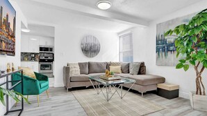 A modern living room with a grey sofa, a glass coffee table, and a green armchair. A large painting and a potted plant are on the walls. - Villa Bonita | 6BR | Pool + Hot Tub (Miami)