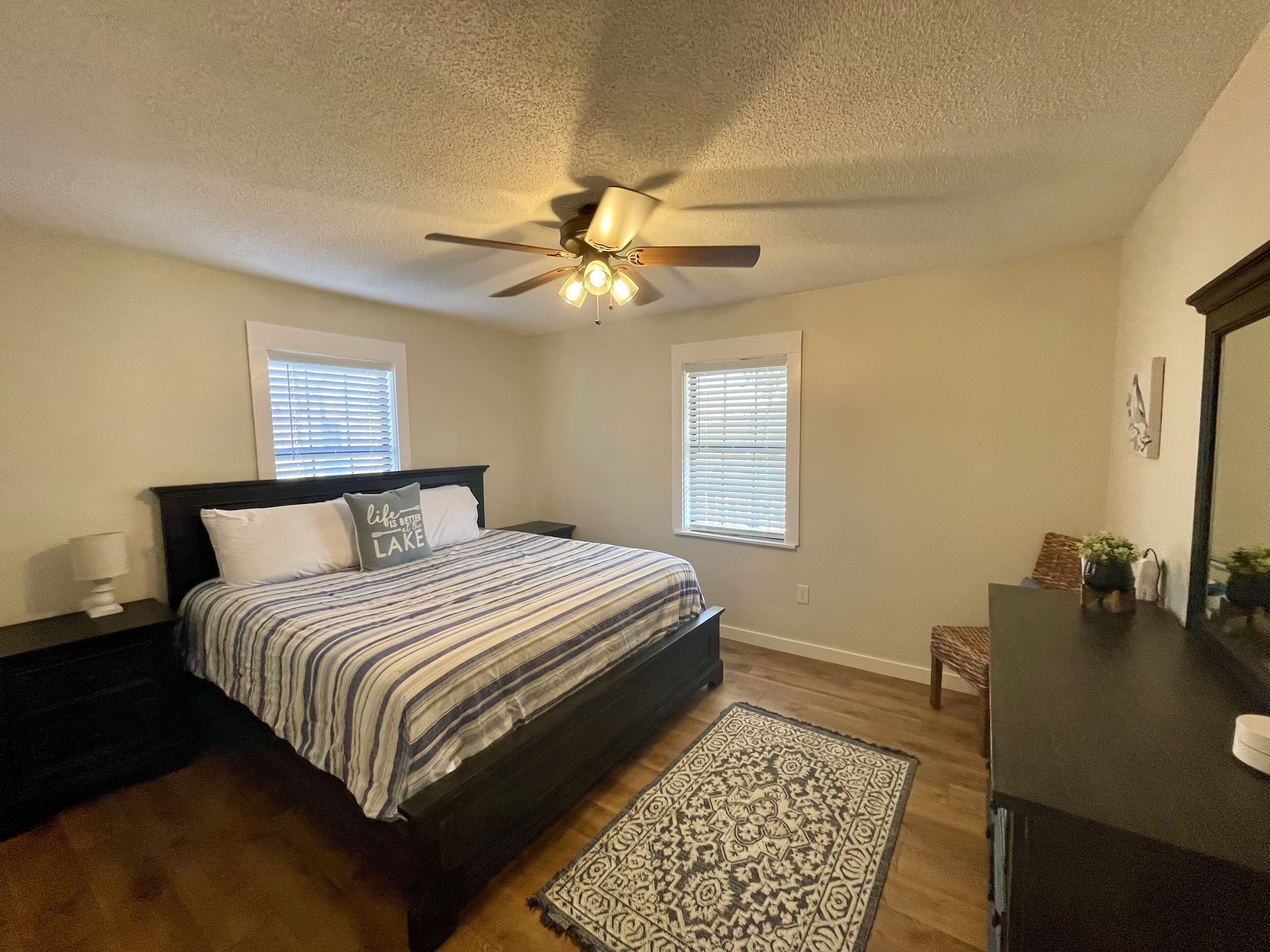 A bedroom with a bed, a ceiling fan, two windows with blinds, a nightstand, a dresser, and a patterned rug.