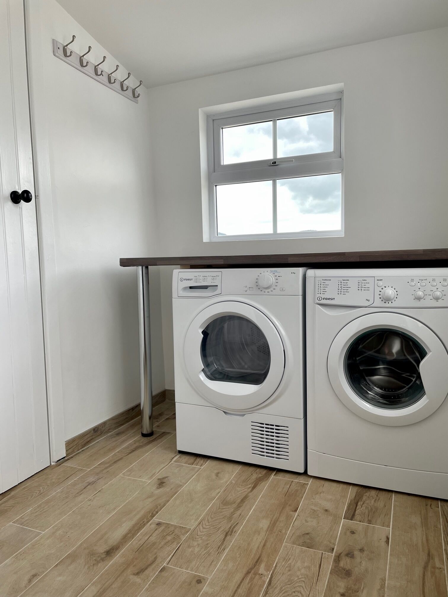 Een wasserette met twee witte wasmachines, een houten vloer, een witte deur en een raam boven de machines.