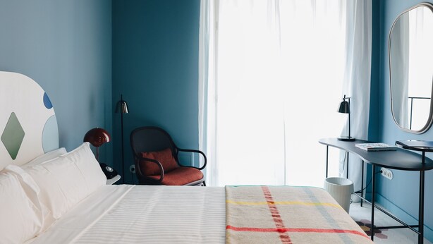 Een slaapkamer met een bed, een stoel, een bureau met spiegel en een raam met gordijnen.