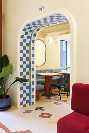 Un hall d’hôtel avec un mur à carreaux, une plante en pot, un fauteuil rouge et un coin repas avec une table et des chaises.