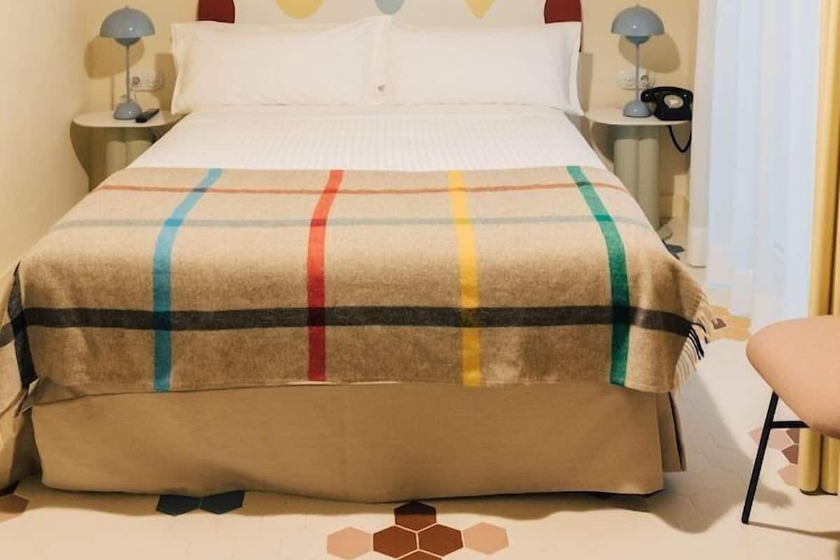 Habitació d'hotel amb llit, taules de nit, làmpada, cadira i finestra amb cortines.