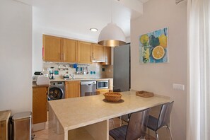 A compact kitchen with wooden cabinets, a built-in dishwasher, and a dining area with a table and chairs. - Apartment "Maré Cheia SL015" with Shared Pool & Wi-Fi (Santa Luzia)
