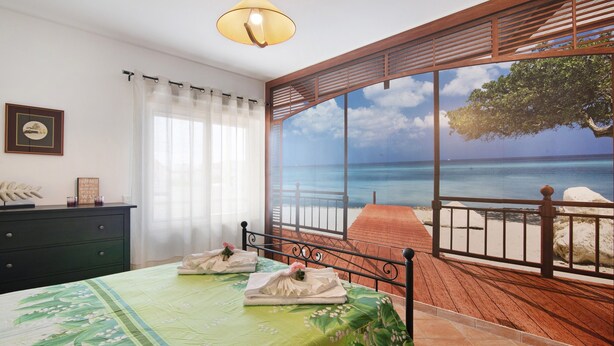 A room with a bed, a view of the ocean, a wooden deck, and a large window with a curtain. - Apartment "Maré Cheia SL015" with Shared Pool & Wi-Fi (Santa Luzia)
