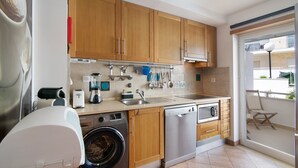 A modern kitchen with wooden cabinets, a dishwasher, a washing machine, and a large window overlooking a balcony. - Apartment "Maré Cheia SL015" with Shared Pool & Wi-Fi (Santa Luzia)