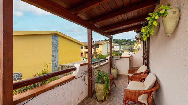 Ein überdachter Balkon mit Korbmöbeln, Blumentöpfen und Blick auf gelbe Gebäude.
