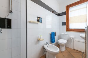 A bathroom with white tiles, a bidet, a toilet, a sink, and a window with blinds.
