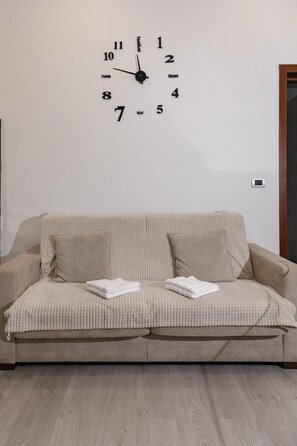 Ein beiges Sofa, eine Uhr an der Wand, ein kleiner Thermostat und ein Holzboden.