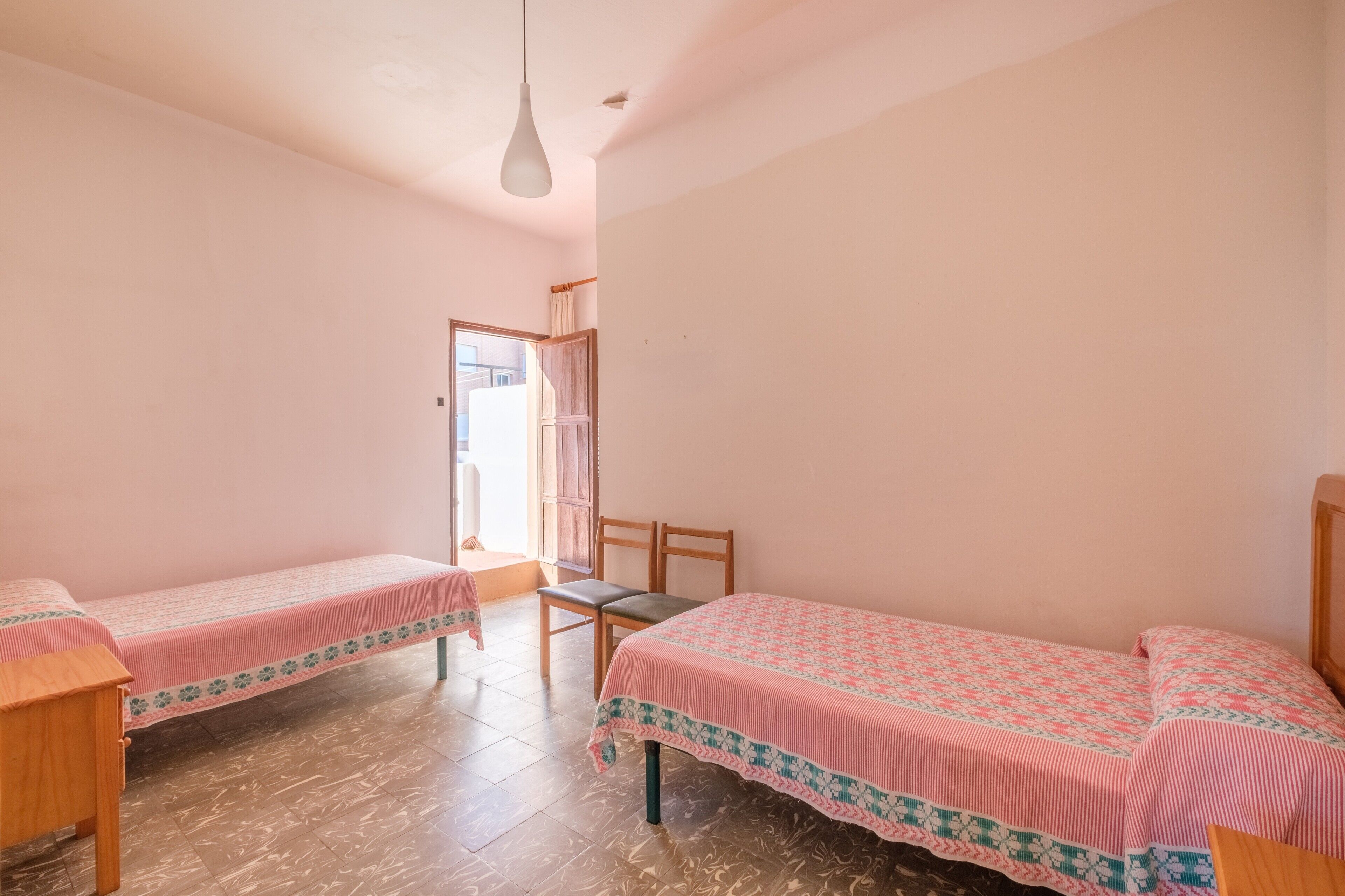 Una habitación con dos camas individuales, una silla de madera, una mesita y una puerta que da a un espacio exterior.