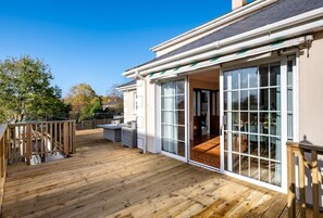 Una terraza de madera con mobiliario exterior, un ventanal de vidrio que da a un espacio interior, y un cielo azul despejado.