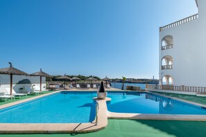 Una piscina con tumbonas y sombrillas, un edificio blanco con arcos y vistas al mar.