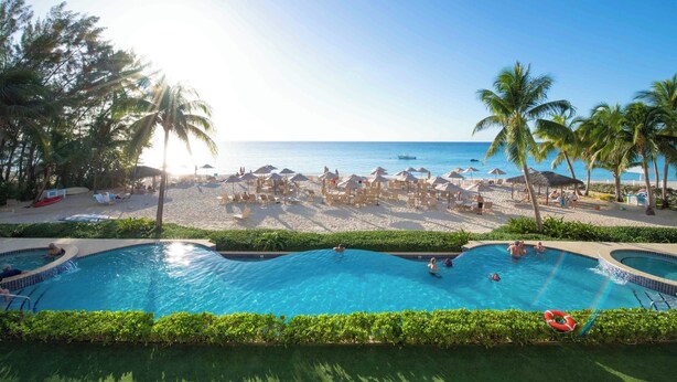 Zona de piscina con agua cristalina azul, rodeada de palmeras y sombrillas de playa.