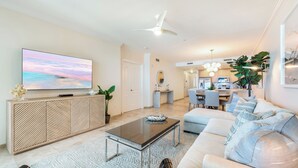 Uma sala de estar moderna com uma grande TV de tela plana, um sofá, uma mesa de centro e uma área de jantar ao fundo.