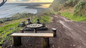 A dirt path leading to a bench with a rustic fire pit, overlooking a coastal landscape with cliffs and a calm sea. - Stylish 2 Bedroom Stone House With Stunning Views, São Jorge, Azores (Vista) (São Jorge)