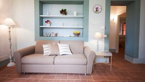 Een woonkamer met een beige bank, een witte bijzettafel met een lamp, en een blauwgroene wandplank met decoratieve objecten.