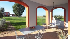 Een overdekt terras met een tafel en stoelen, een hanglamp en uitzicht op een groen landschap en een rood gebouw op de achtergrond.