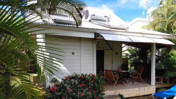 Una casa pequeña con un porche cubierto, un jardín con flores rojas y una silla lounge azul.