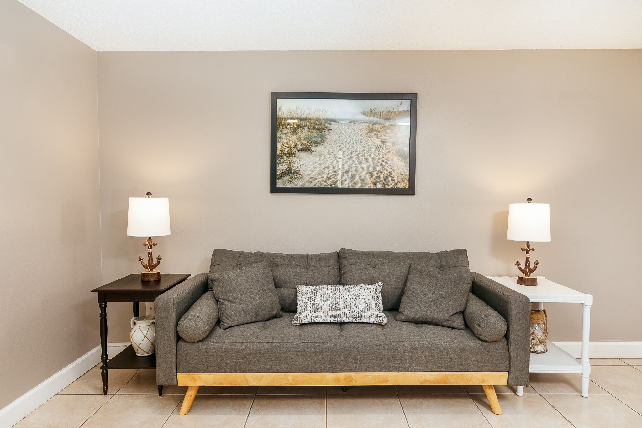 A living room with a grey sofa, two small side tables with lamps, a framed beach picture on the wall, and a patterned throw pillow.