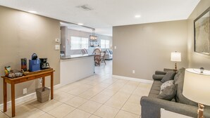 A spacious living room with a gray sofa, a wooden coffee table, and a lamp. A kitchen area with a dining table and chairs is visible in the background.