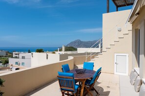Una terraza con una mesa y sillas, con vista al mar y edificios, y una montaña al fondo.