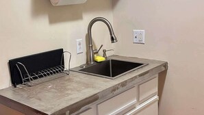A small kitchenette with a sink, a countertop, a shelf above the sink, and a roll of paper towels.