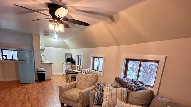 A spacious living area with a sofa, armchair, and coffee table. A ceiling fan and kitchen are visible in the background.