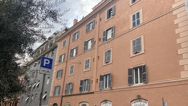 Un immeuble à plusieurs étages, avec un panneau indiquant un parking, des voitures garées et un café orné d’une fresque murale.