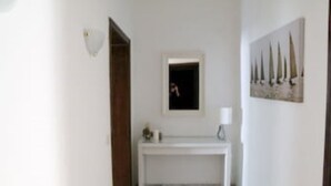 Un couloir avec une console blanche, un tableau encadré représentant des voiliers et une porte donnant sur une autre pièce.