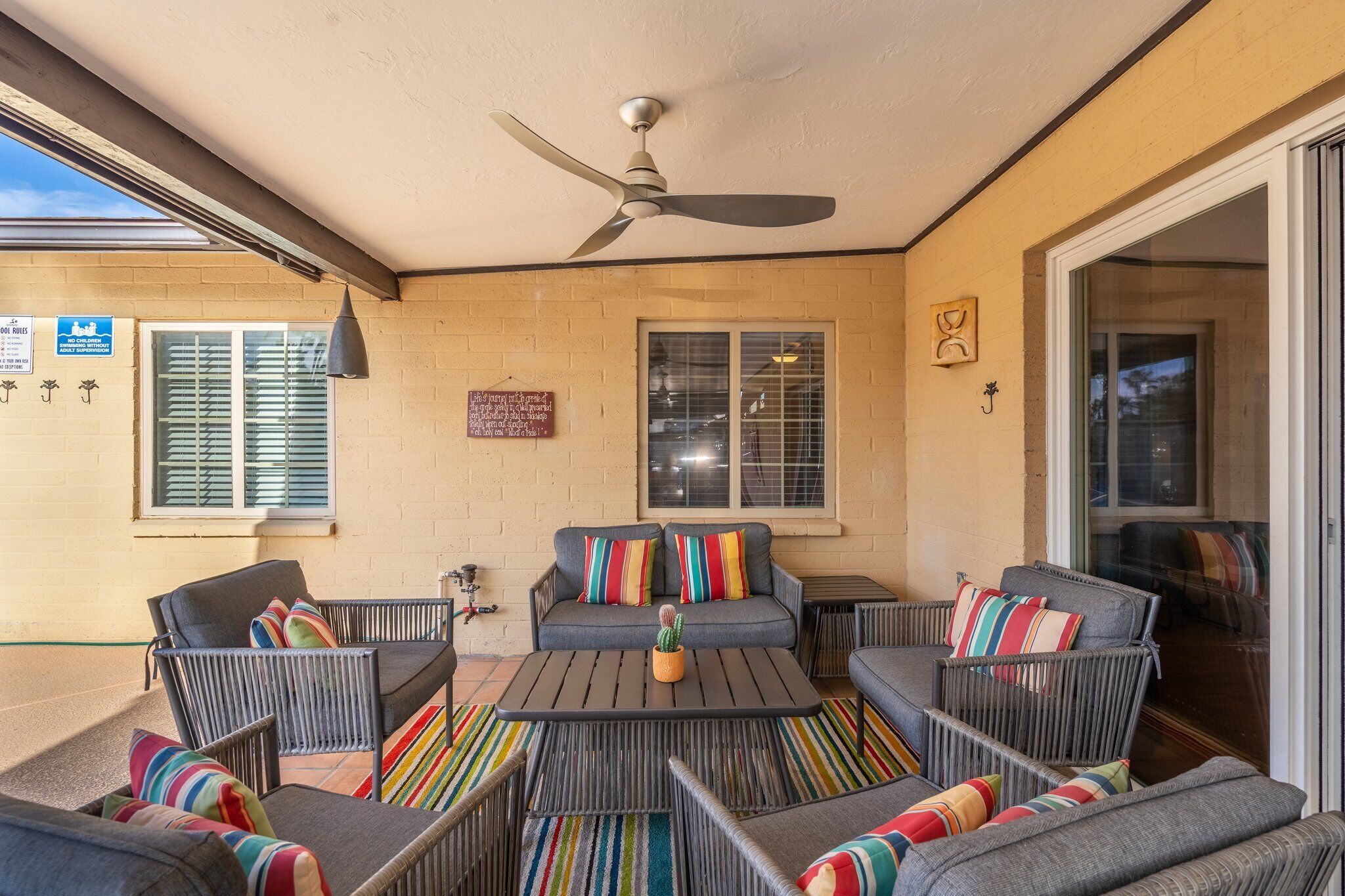 Un patio techado con ventilador de techo, un conjunto de sofás grises con cojines de colores, una mesita con un cactus y una ventana con persianas.