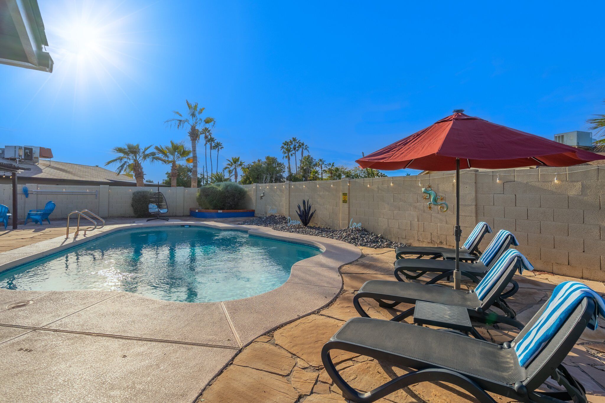 A swimming pool with lounge chairs and an umbrella, surrounded by palm trees and a brick wall.