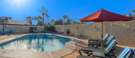 A swimming pool with lounge chairs and an umbrella, surrounded by palm trees and a brick wall.
