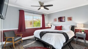 Ein Schlafzimmer mit Bett, Sessel, Nachttisch, Deckenventilator und einem Fenster mit Vorhängen.
