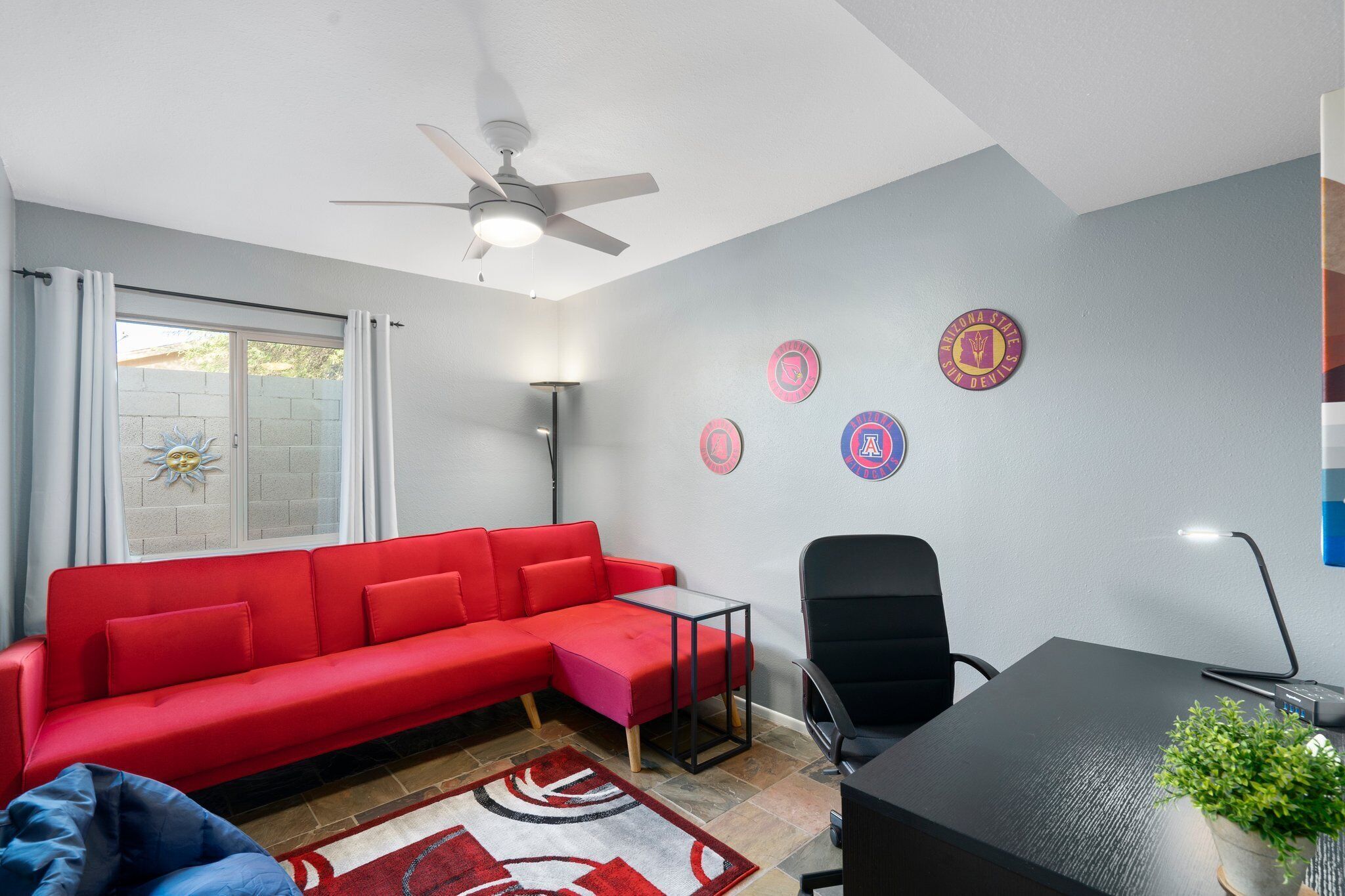 A room with a red sofa, a black office chair, a desk with a lamp, a ceiling fan, and a window with curtains.