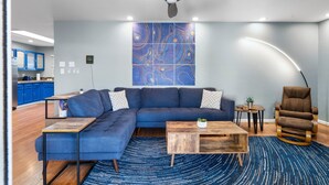 Ein modernes Wohnzimmer mit einer Eckcouch, einem Holztisch, einem blauen Teppich mit Muster sowie einem Deckenventilator. Im Hintergrund ist eine Küchenzeile erkennbar.
