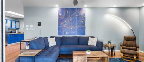 A modern living room with a sectional sofa, a wooden coffee table, a blue patterned rug, a ceiling fan, and a kitchen area visible in the background.