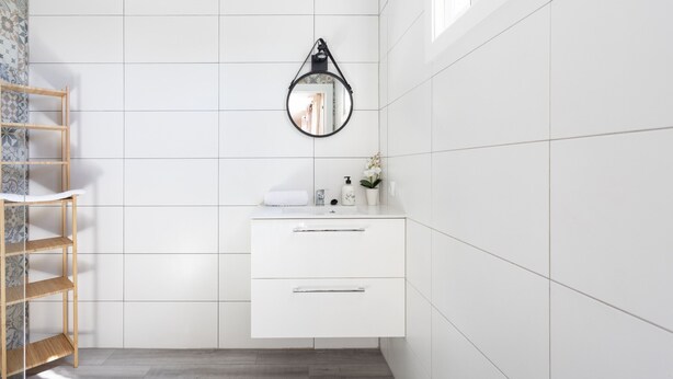 Une salle de bain avec des murs carrelés de carreaux blancs, une étagère en bois, un miroir rond et un meuble-lavabo blanc avec des tiroirs.
