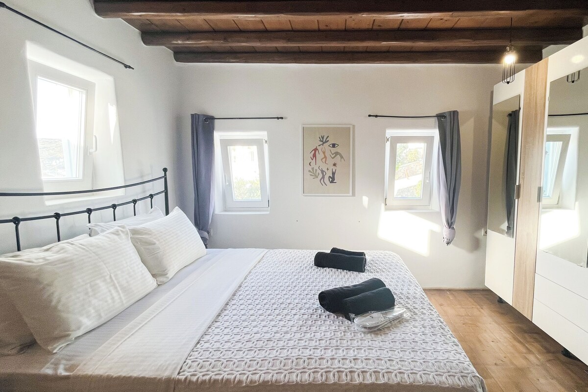 A bedroom with a bed, two windows, a mirror, and a wall with a picture.